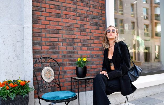 Fashionable woman in a black suit and sunglasses poses against a brick wall in Tallinn's urban setting.