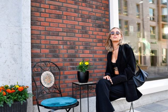 Fashionable woman in a black suit and sunglasses poses against a brick wall in Tallinn's urban setting.